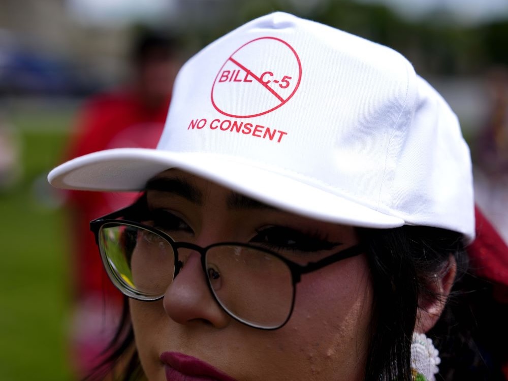 Close-up of a person with medium-light skin and glasses wearing a white ball cap with the words ‘Bill C-5’ in a crossed-out circle and ‘No Consent’ in red lettering.