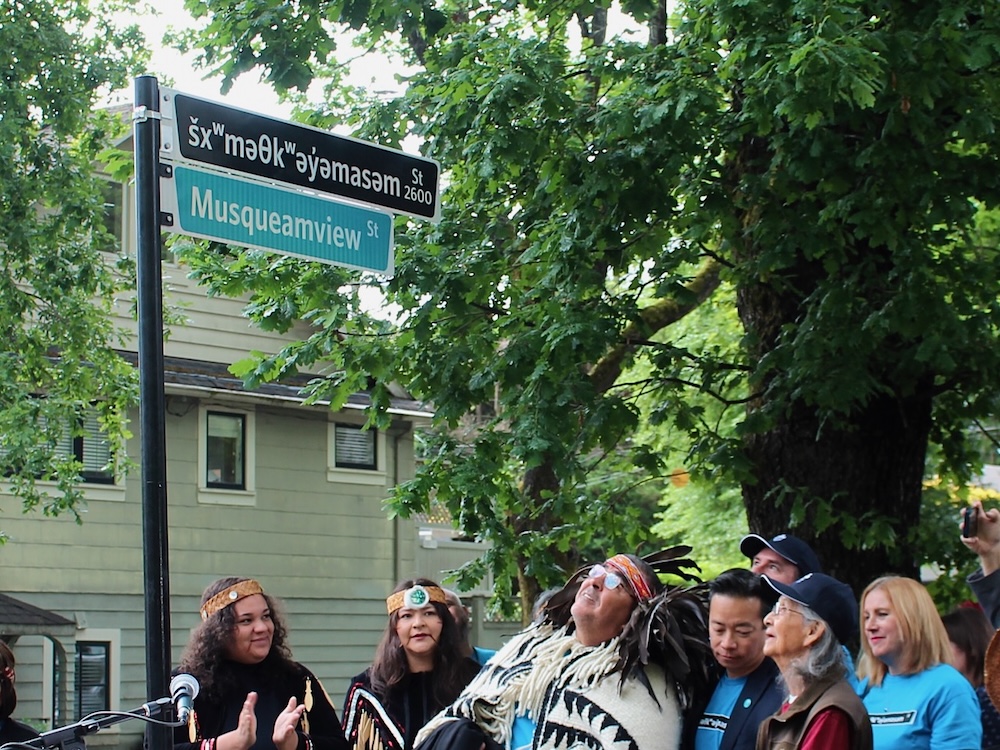 A man wearing glasses and Musqueam regalia looks up at a street sign that says ‘šxʷməθkʷəy̓əmasəm Street.’ To the left of the frame, two women wearing cedar headbands and black clothing decorated with wooden paddles applaud. To the right, more people are gathered, most looking up at the street sign with happy expressions on their faces.