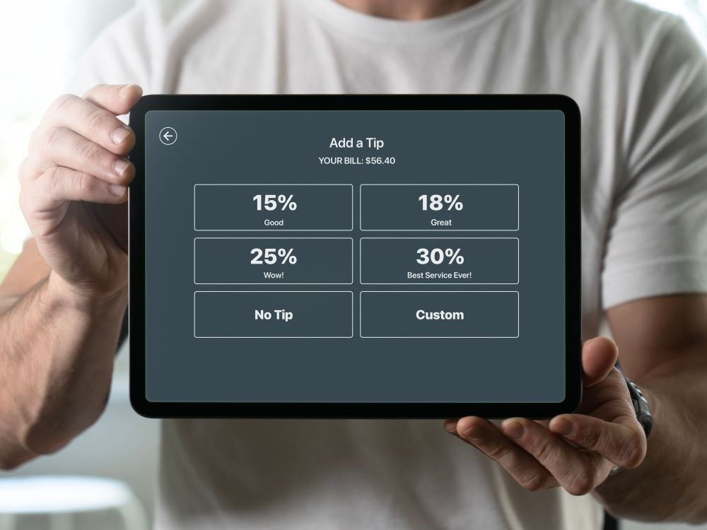 A close-up shows a computer payment screen with suggested tips, held in the hands of a person.