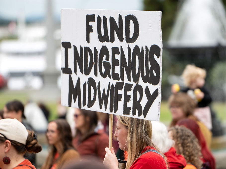 A woman with long blond hair holds a white sign with black letters that read ‘Fund Indigenous midwifery.’