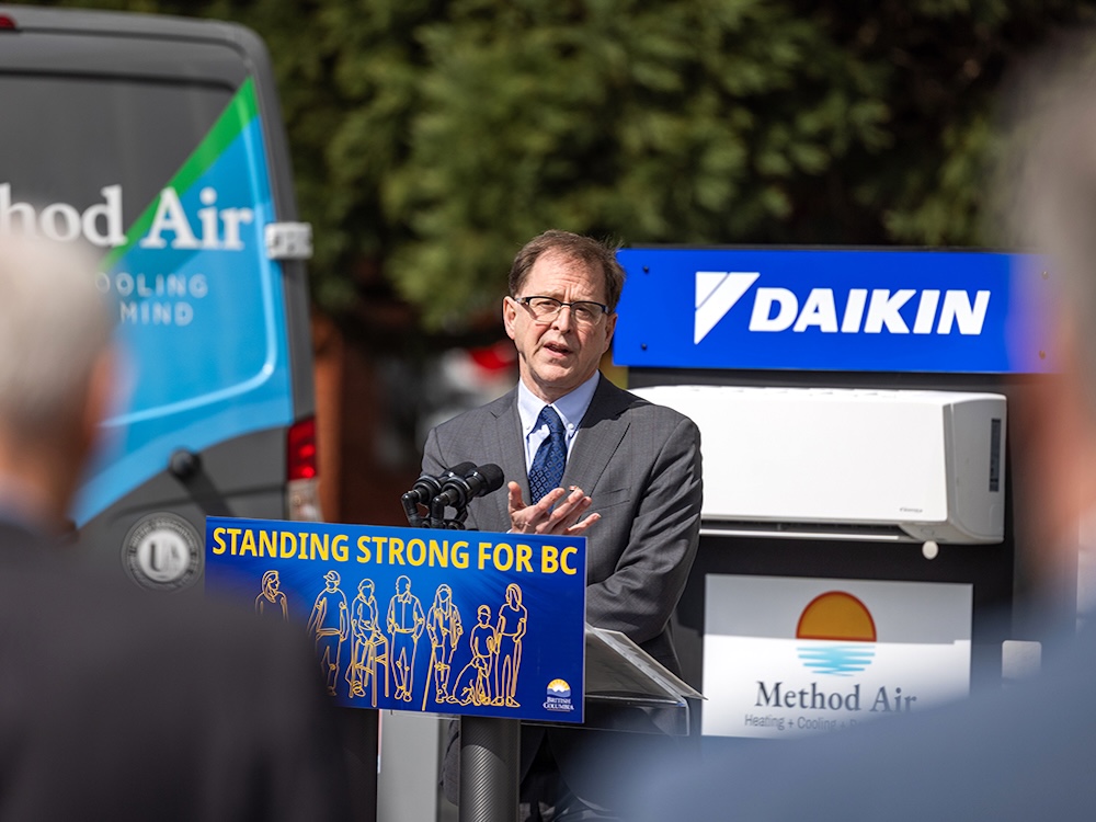 A man with short brown hair and glasses stands at a podium that says ‘Standing Strong for BC.’