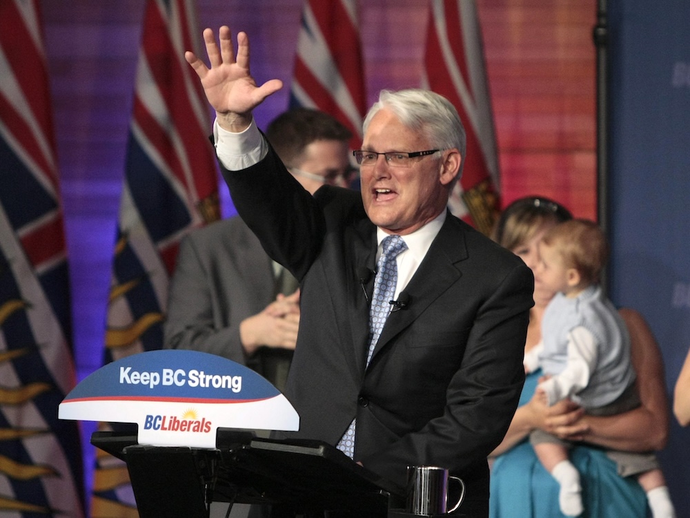 A 60-year-old light-skinned man with grey hair, wearing a dark suit, waves in celebration on an election-night stage.