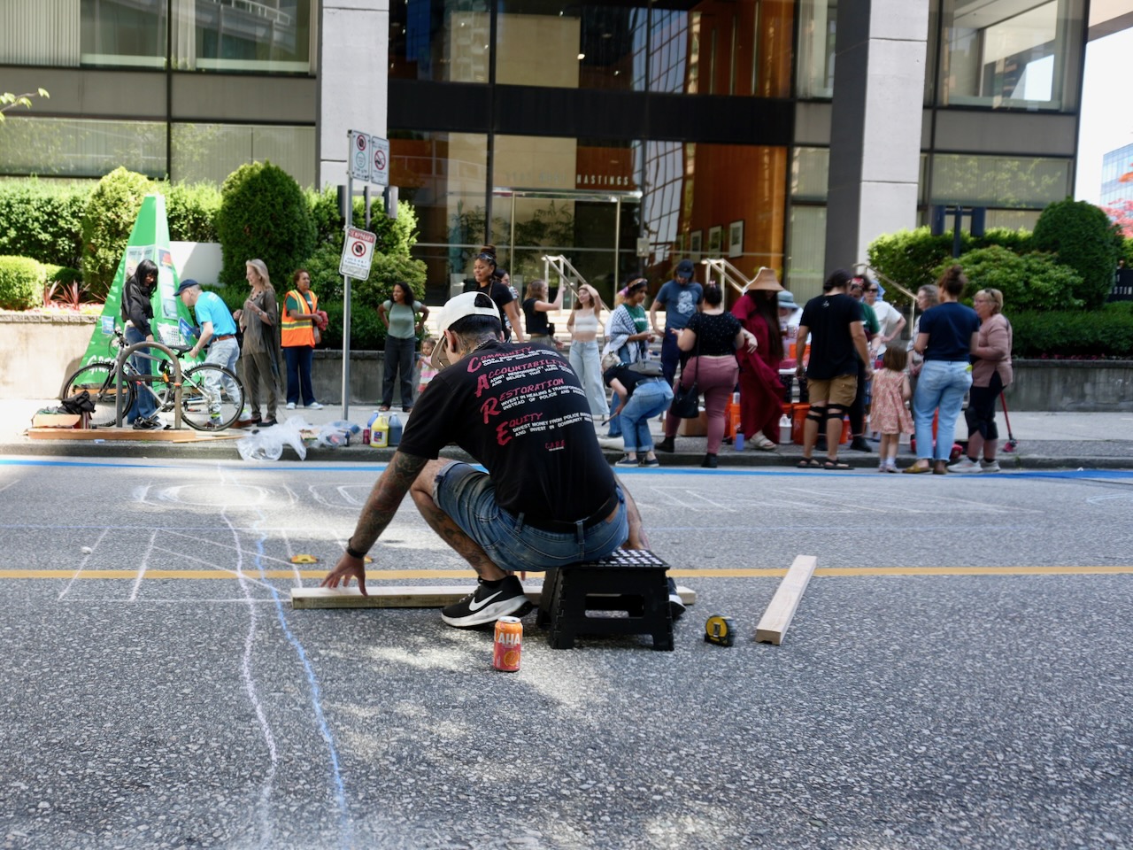 A man in a black baseball cap and a black t-shirt crouches over a sidewalk, measuring and using chalk to draw. In the background a group of people can be seen.