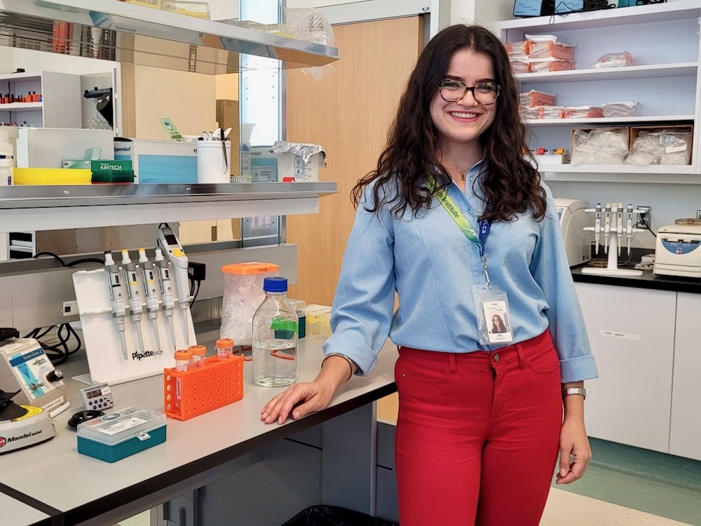 A woman with long curly brown hair grins at the camera as she stands in her laboratory, which has grey counters and several shelves with brightly-coloured scientific equipment.
