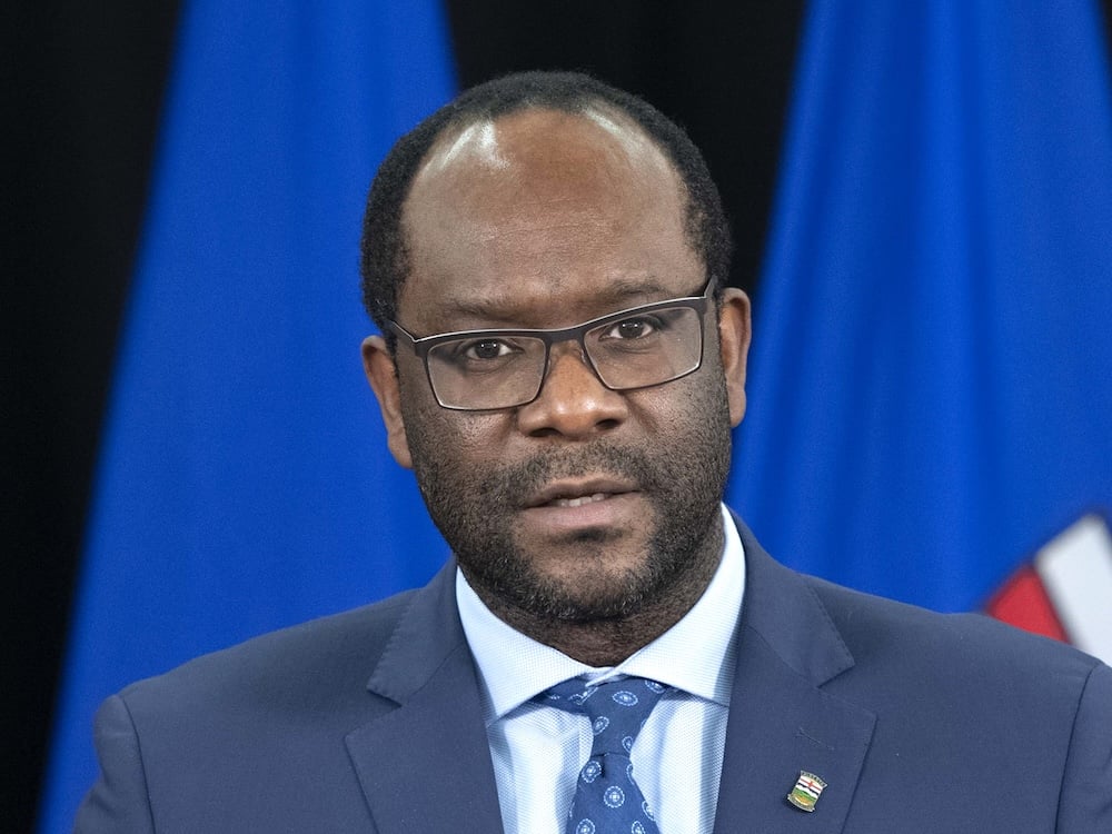 A 50-ish Black man looks at the camera. He has short, receding black hair and stubble and wears glasses, a blue suit, a whitish shirt and tie.