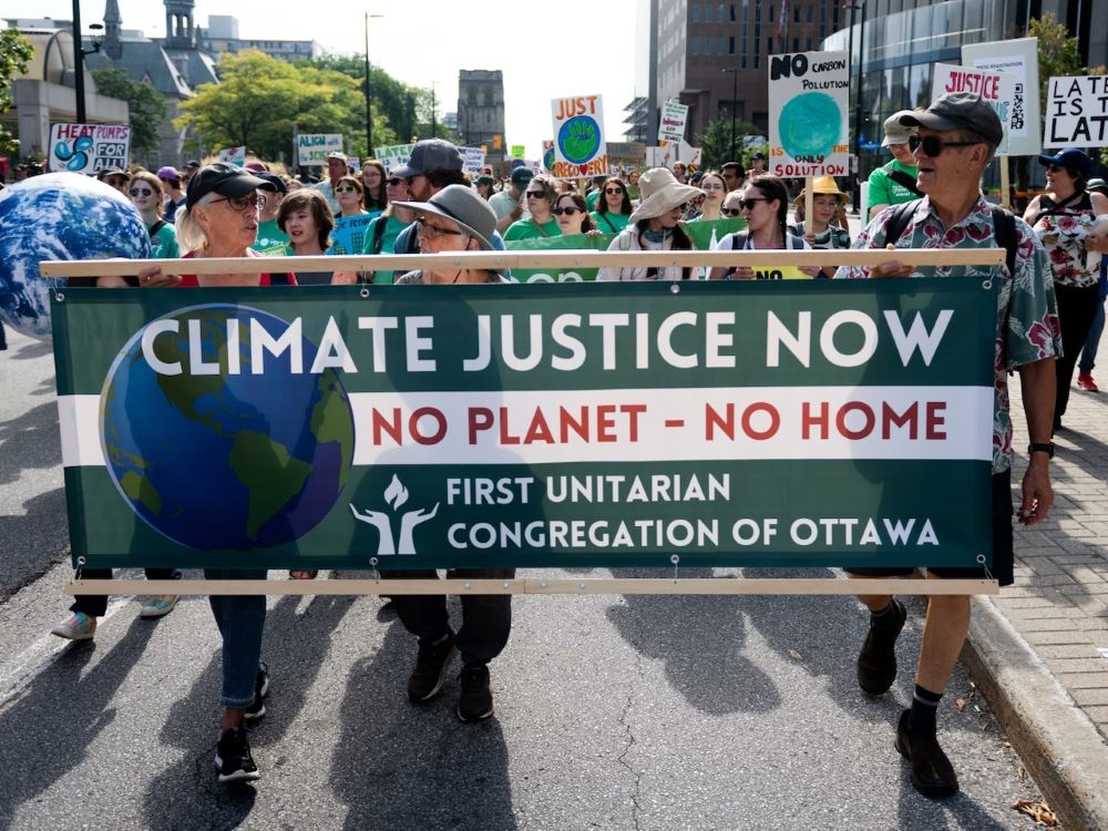A group of people march in the road on a sunny day, holding a banner that reads “Climate Justice Now.”