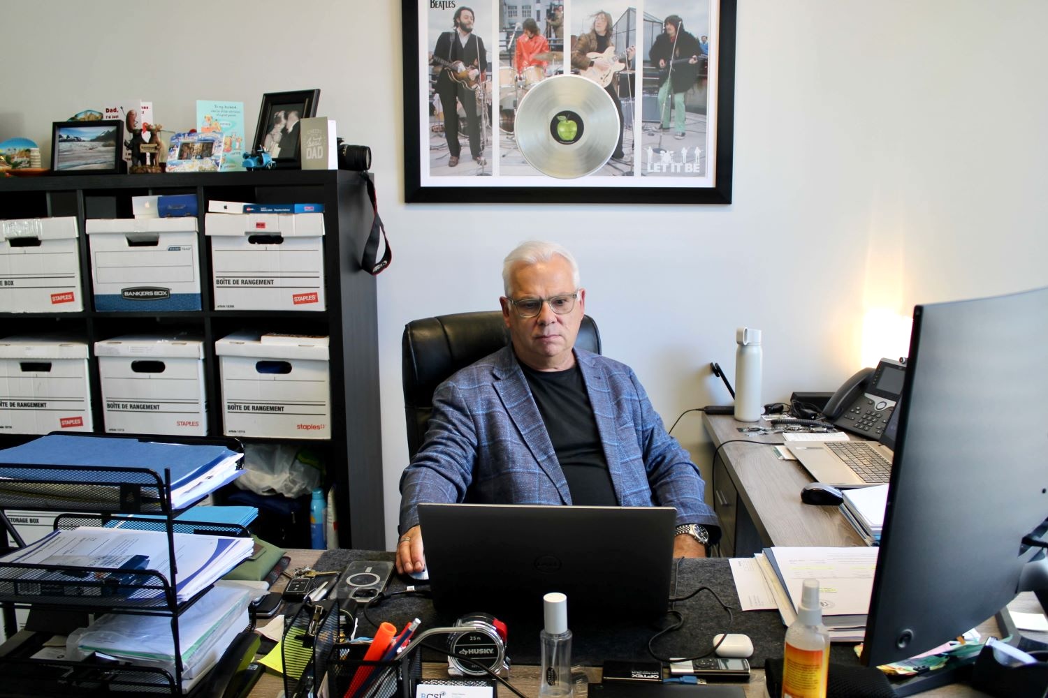 A light-skinned middle-aged man with white hair sits at a desk with boxes of files behind him. He wears a black T-shirt and a blue sports jacket.