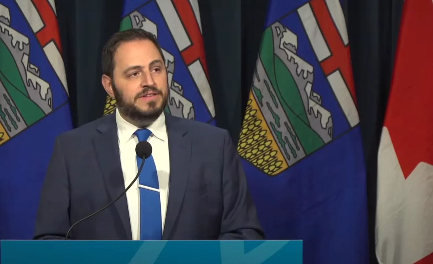 A light-skinned man in his 30s with dark hair and beard and wearing a suit stands at a podium.