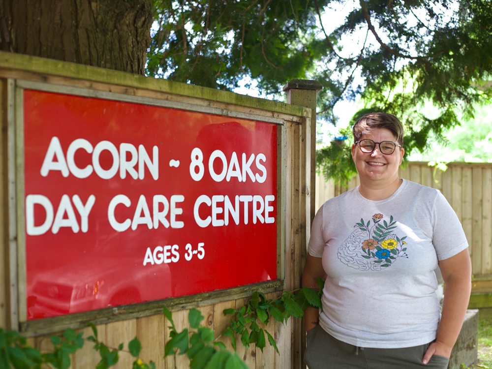 A person with short brown hair and light skin tone wears a T-shirt and glasses and stands in front of a sign that reads, &lsquo;Acorn 8 Oaks Day Care Centre, Ages 3 to 5.&rsquo;
