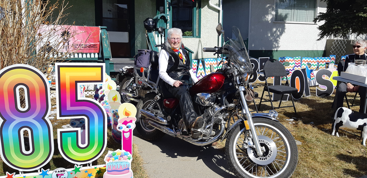 An older woman with a silver mullet and light skin tone sits on a motorcycle. In the foreground is a sign that reads, ‘85,’ with birthday candles.