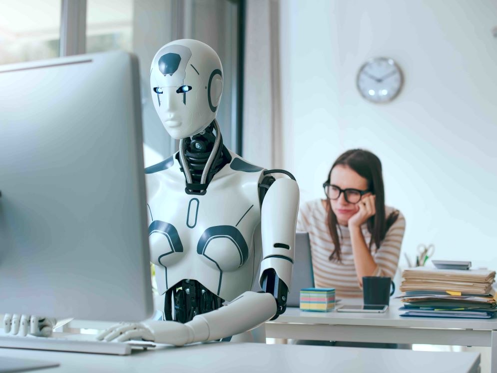 A robot sits at a desk working on a computer, with a discouraged-looking woman at a similar workstation behind it.