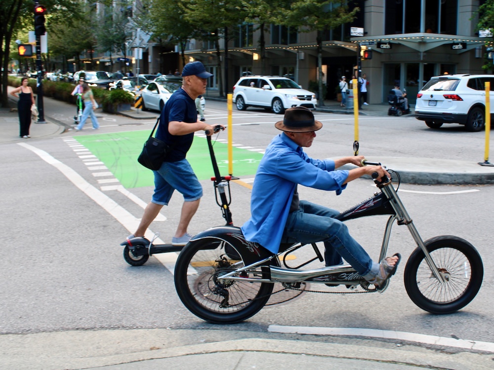 A street scene showing a man wearing a fedora riding an e-bike and another man wearing a ball cap standing riding a scooter.