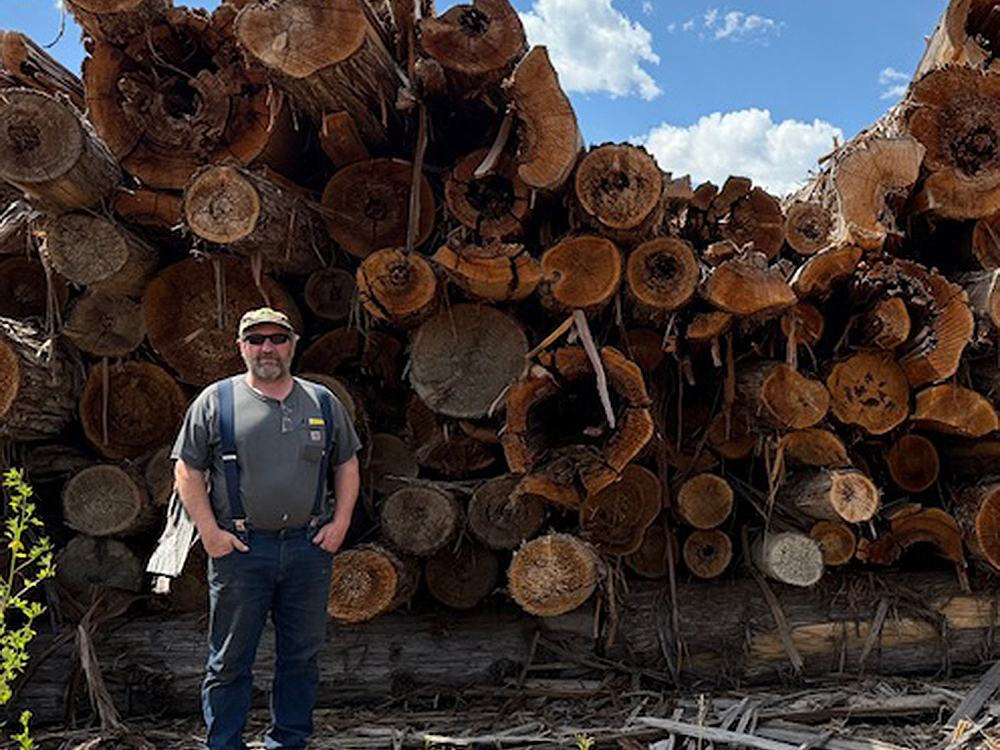A man with light skin tone wears a ball cap, sunglasses, T-shirt, suspenders and dark pants. He is standing in front of a pile of cedar logs.