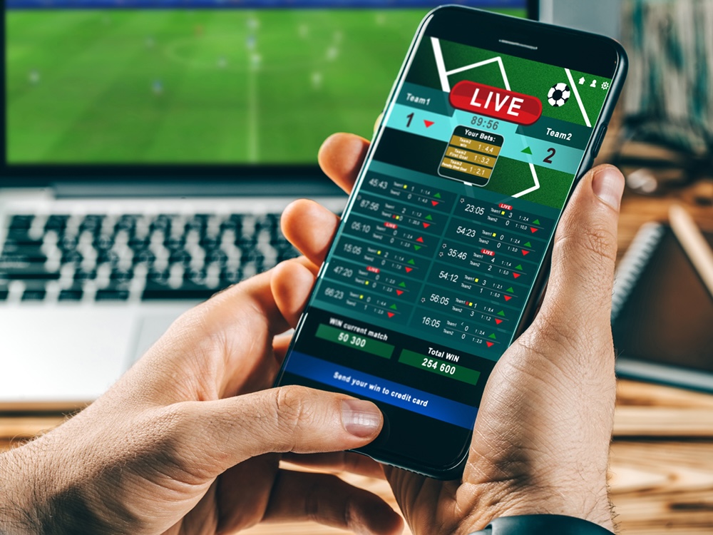 Close-up cropped shot of male hands making bets using a mobile application on his phone. He is watching a soccer game on his laptop.