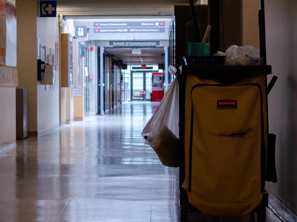 A hallway at a university. In the foreground, a cleaning trolley can be seen with a large yellow bag and cleaning supplies.