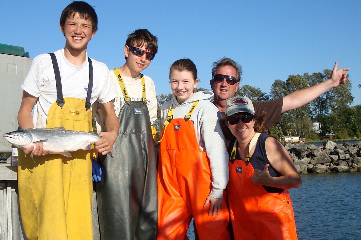 Five people, including two adults and three adolescents, stand in front of a large water body and smile. They are wearing brightly covered overalls. A boy on the left holds a fish.
