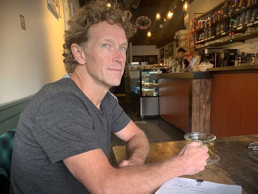 Author J.B. MacKinnon sits at a table in a caf&eacute; with a cup of tea in front of him. He has light skin tone, blue eyes and curly blond hair and is wearing a dark grey T-shirt.