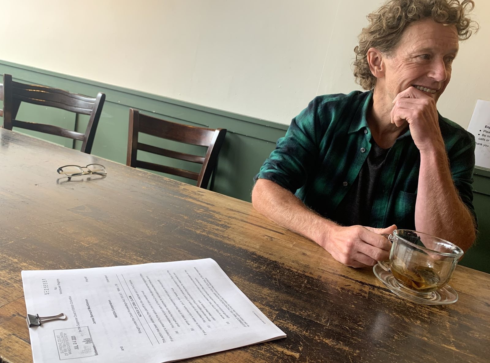 J.B. MacKinnon sits at a table with a clipped sheaf of legal papers in front of him. He has light skin tone, blue eyes and curly blond hair and is looking toward the right of the frame.