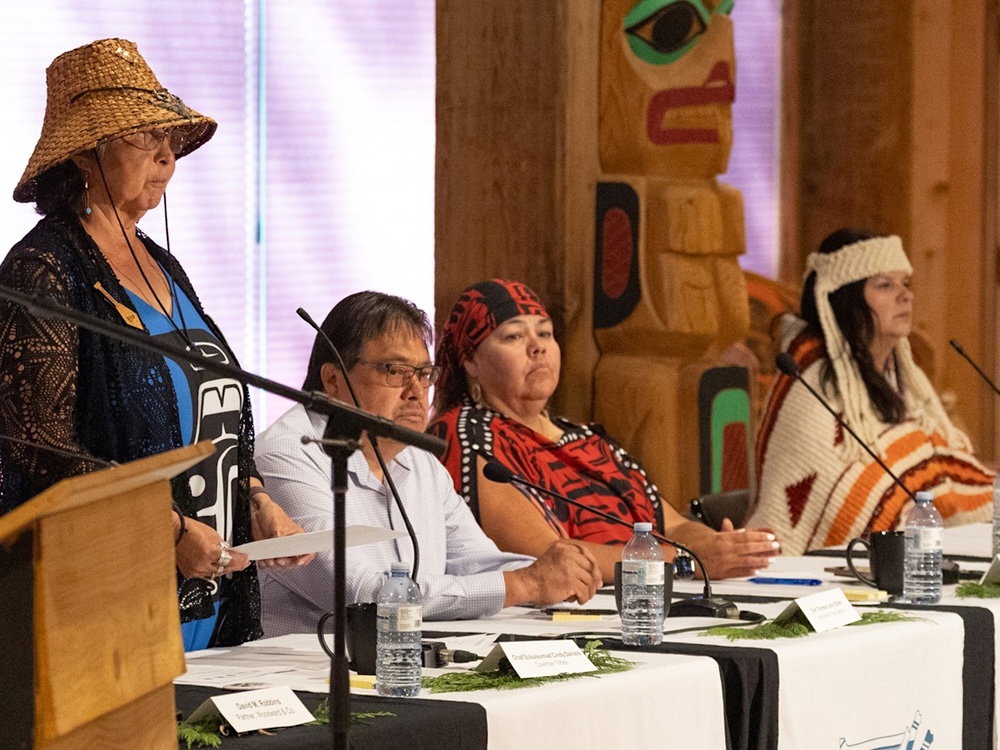 Chief Sulsulxumaat Cindy Daniels of Cowichan Tribes stands and speaks. She is wearing a traditional woven cedar hat and a blue shirt with Coastal First Nations art.
