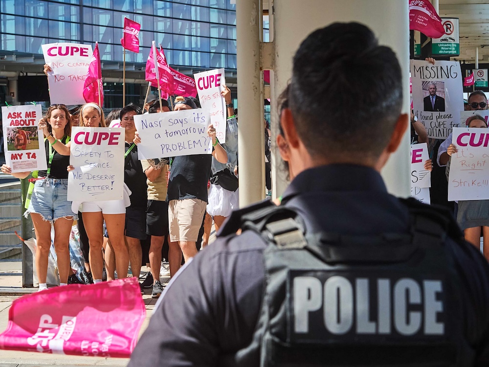 A group of picketers stand in the sun holding signs. In the foreground, a uniformed police officer watches them.