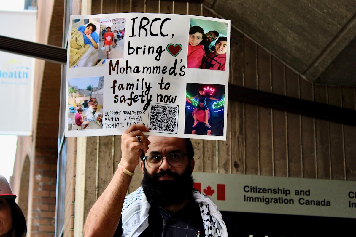 A man with medium skin tone and a long black beard holds a poster showing pictures of his family.