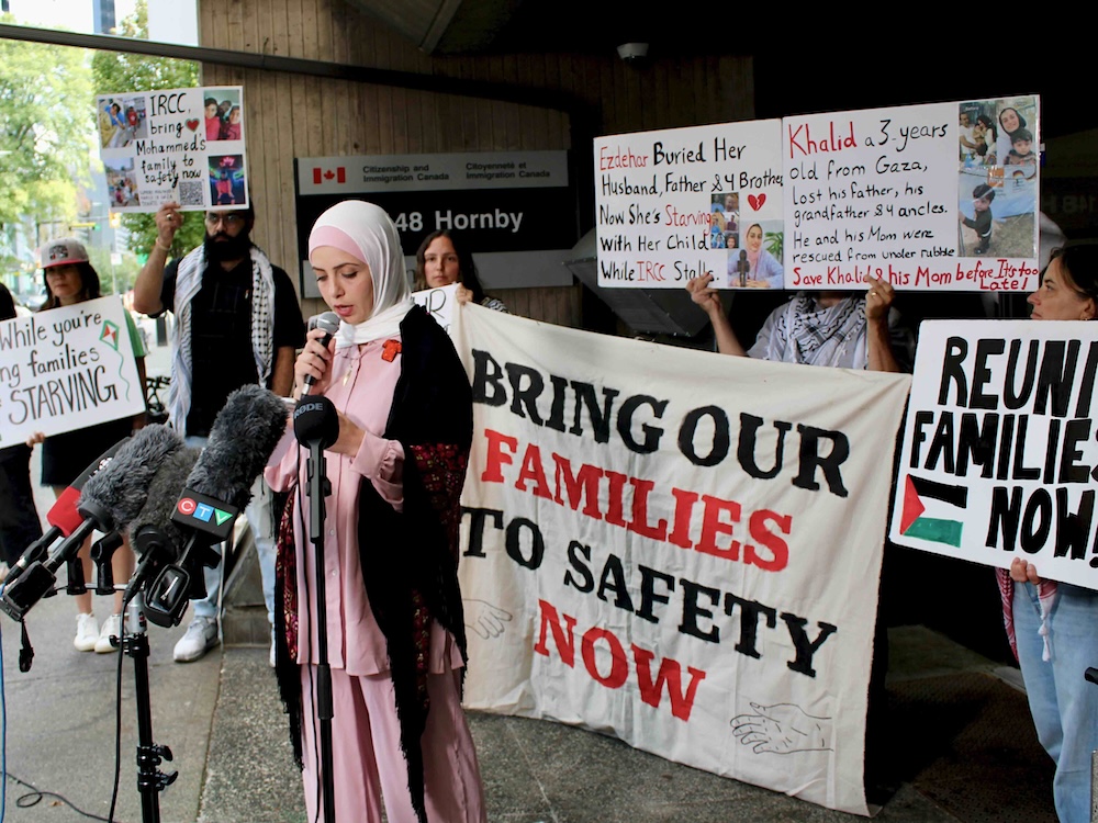 A woman wearing a pink and white headscarf and a pink shirt and pants holds a microphone. Behind her, supporters hold signs calling for Gaza families to be reunited in Canada.