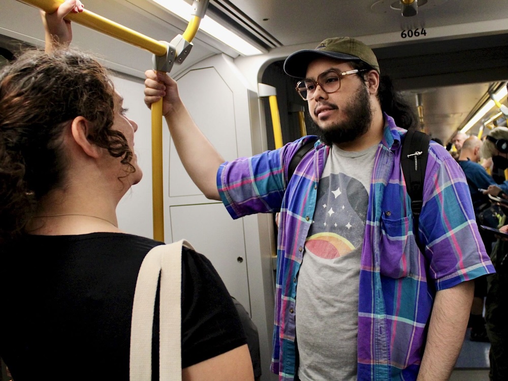 A man with long brown hair in a ponytail, wearing a light grey ball cap and glasses, speaks to a woman with brown curly hair in a ponytail as they both hold on to poles while riding in a SkyTrain car.