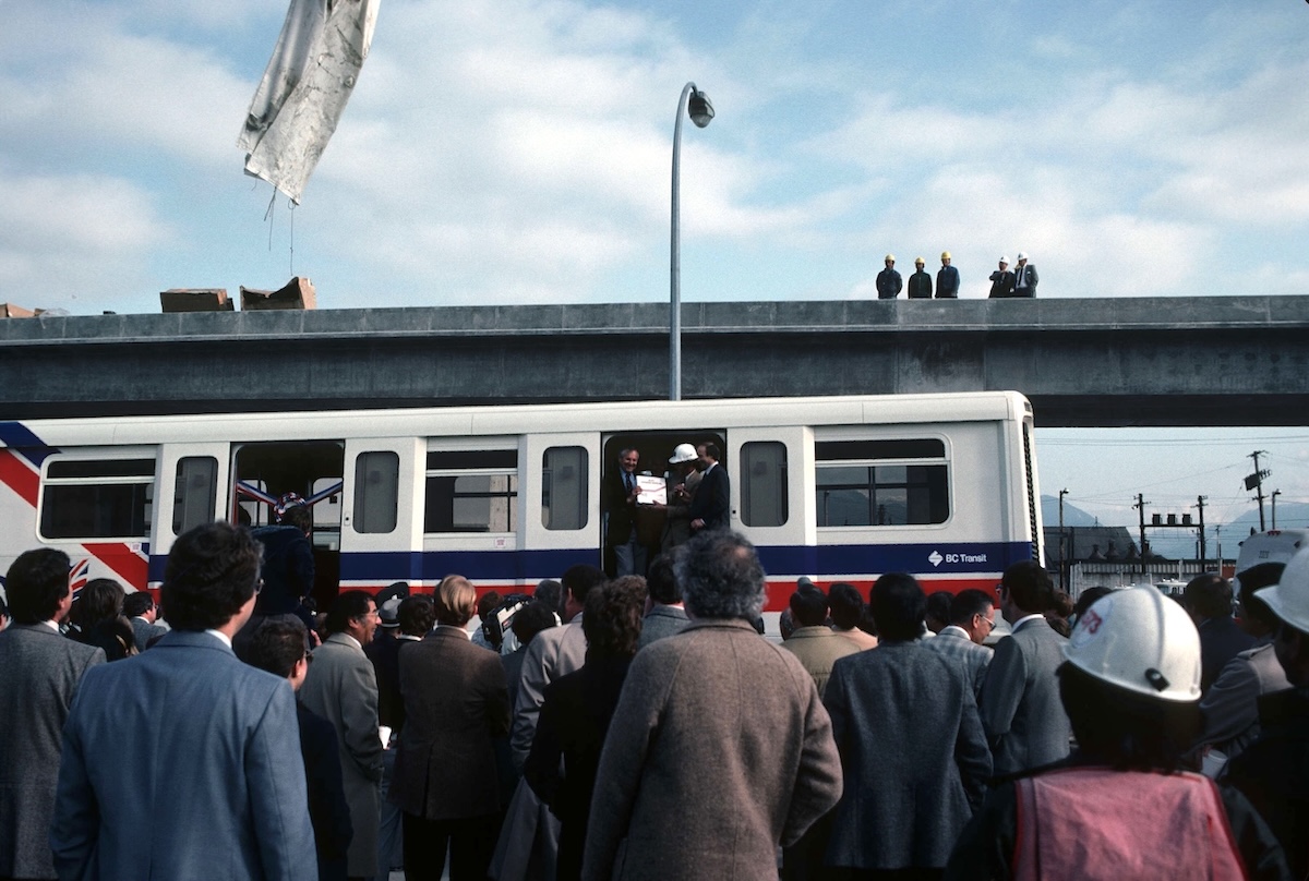 A crowd of people stand before a train with a ribbon over one door and two people holding up a plaque in another door. Five people in hard hats look down on the scene from an elevated concrete track above and behind the train.