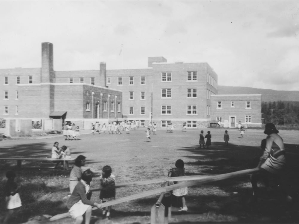 An old black and white photo of a four-storey institution, with children visible in the yard in front. Some sit on a pair of see-saws in the foreground.