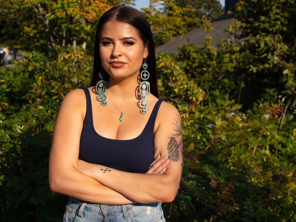 A woman in her 20s with long dark hair wearing a tank top and with dangling turquoise earrings stands outdoors with arms folded.