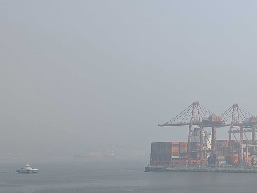 A city port with cranes is visible through a hazy grey cloud of smoke. The SeaBus is visible in the foreground.