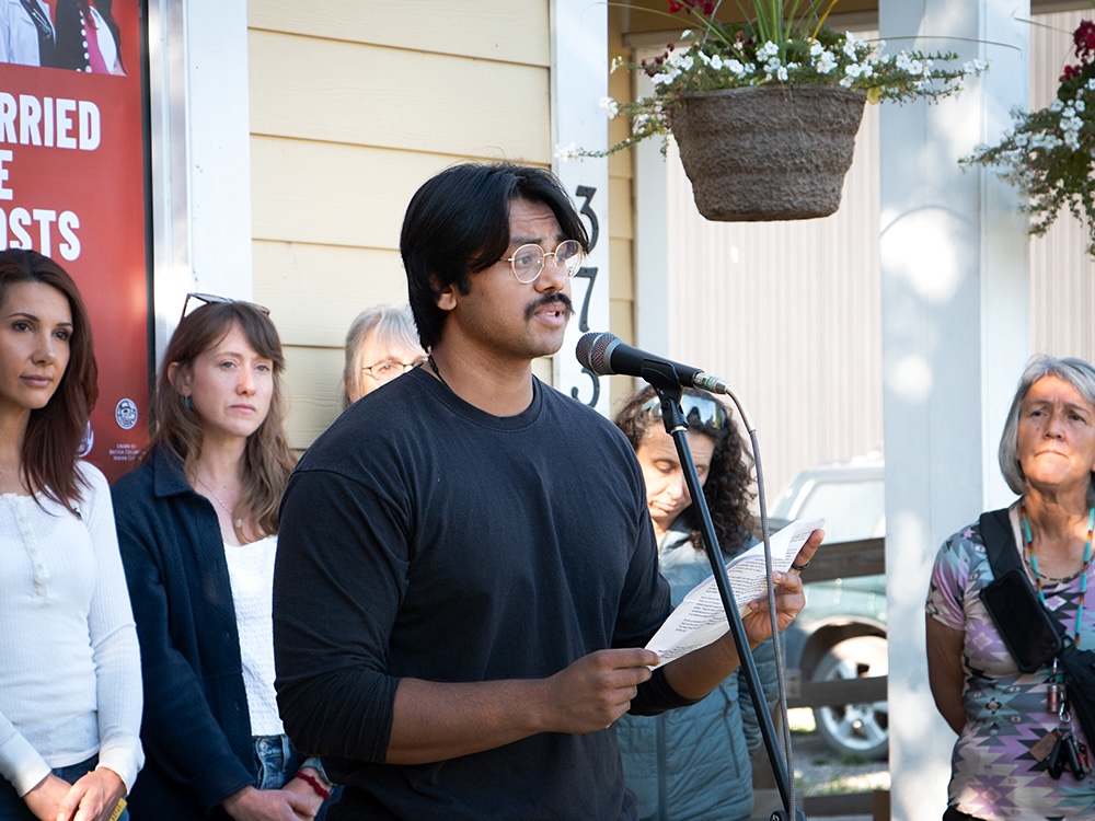 A man with medium-dark skin tone, dark hair and moustache, speaks into a microphone. Five people stand behind him.