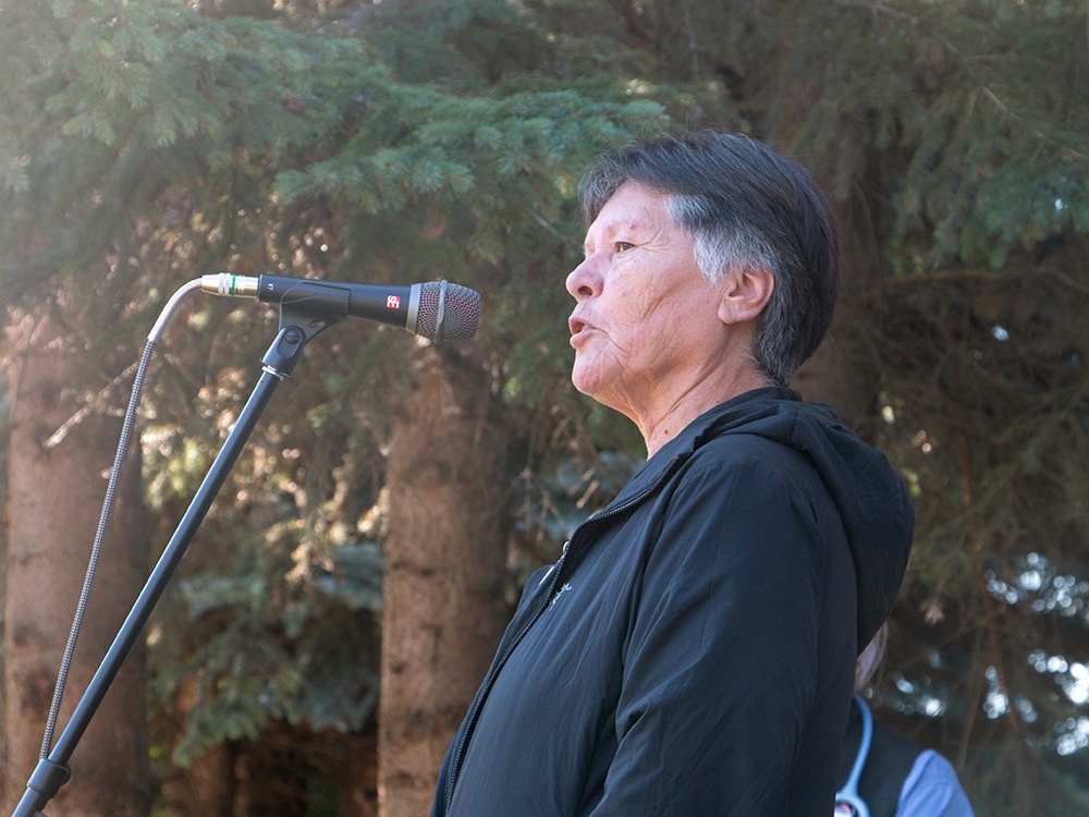 A woman with short, dark hair and wearing a black jacket speaks into a microphone. There are trees behind her. 