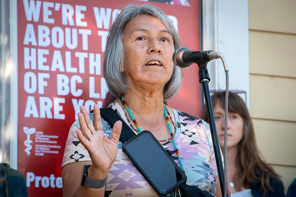 A middle-aged woman with grey hair stands at a microphone.