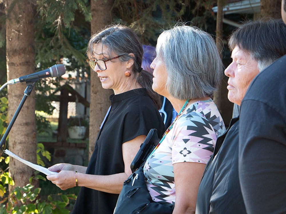 Three women stand in an outdoor setting with trees behind. The woman on the left, who has dark hair and wears glasses, speaks into a microphone.