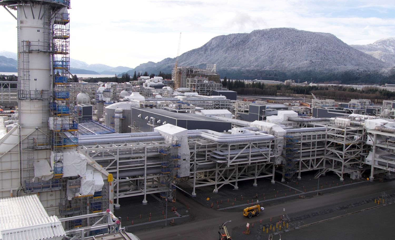 A sprawling complex of pipes and buildings is in front of a stretch of water and a mountain.