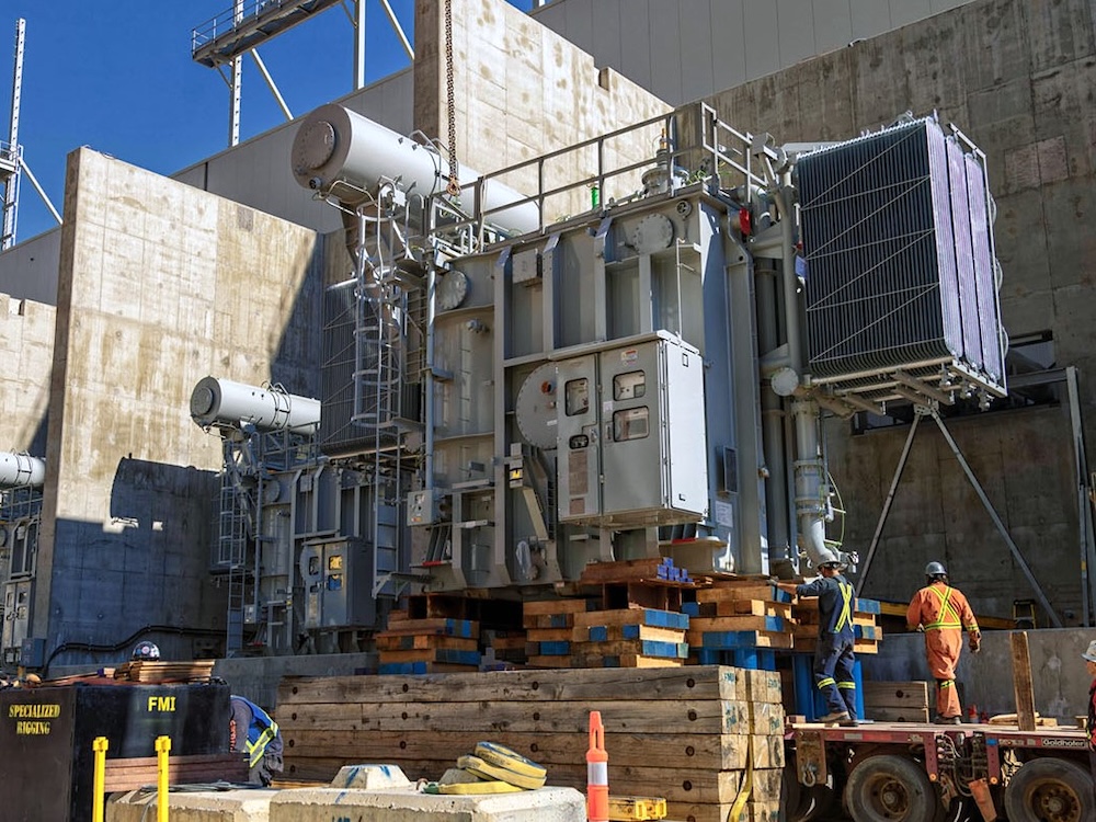 Workers in red safety suits and helmets are in front of a large concrete structure.