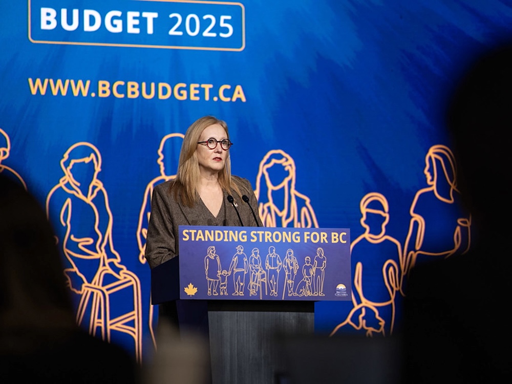 Brenda Bailey, a 40-ish person with blond hair, stands at a lectern with a background that says &lsquo;Budget 2025.&rsquo;