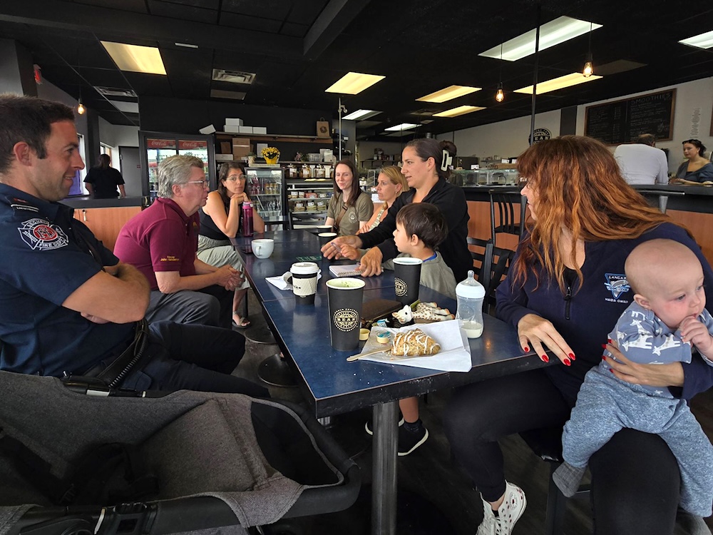 A light-skinned man in a firefighter&rsquo;s navy uniform and a light-skinned, grey-haired man in a maroon polo shirt sit across a table from a brown-haired woman in a green shirt, a blond woman with short hair, a brown-haired woman holding a notebook, a light-skinned brown-haired boy and a red-haired woman holding a baby. A bottle and toy giraffe sit on the table amid coffee cups.