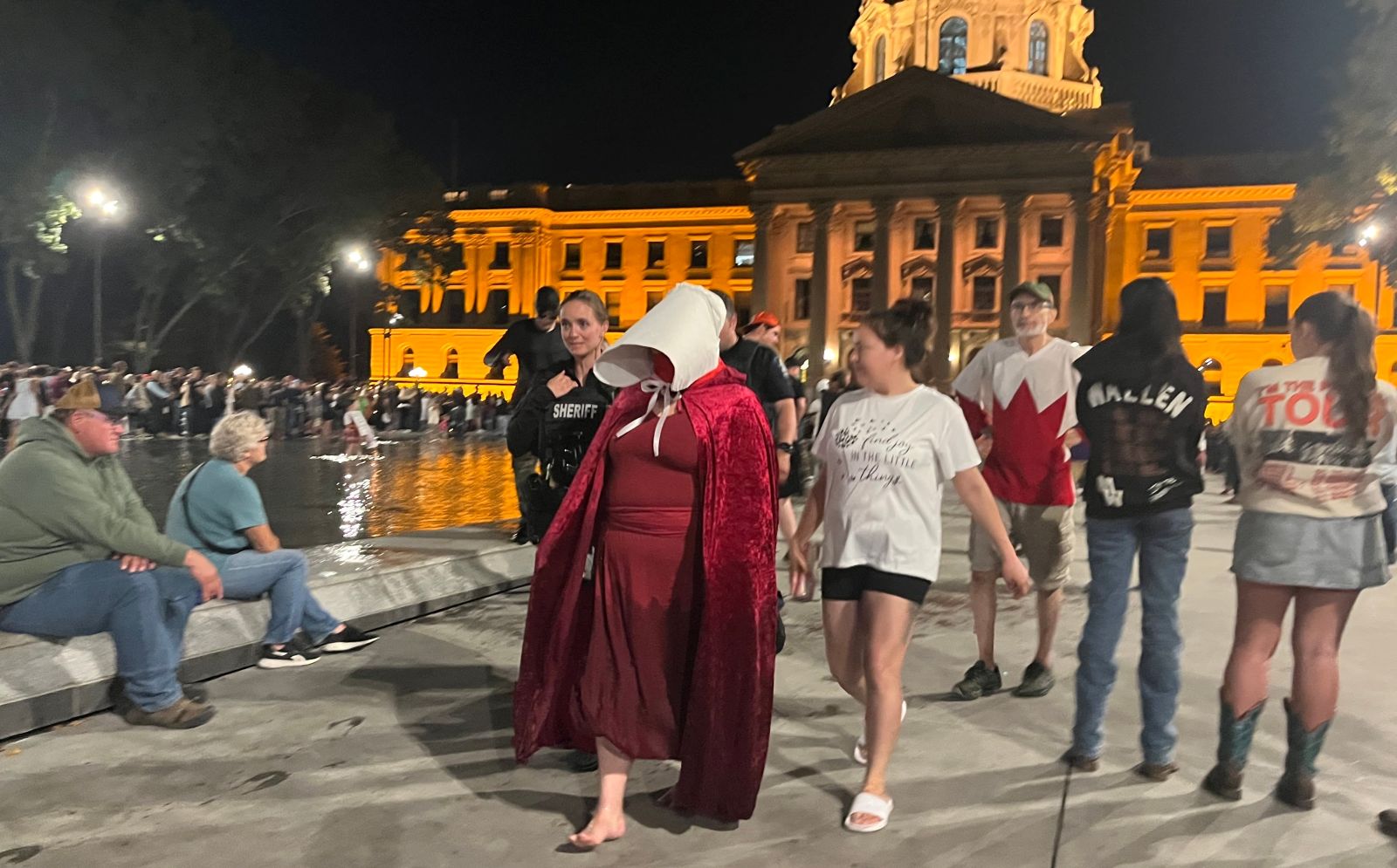 A person walks barefoot, wearing red robes and a white headdress that hides her face, in front of the stone Alberta legislature.