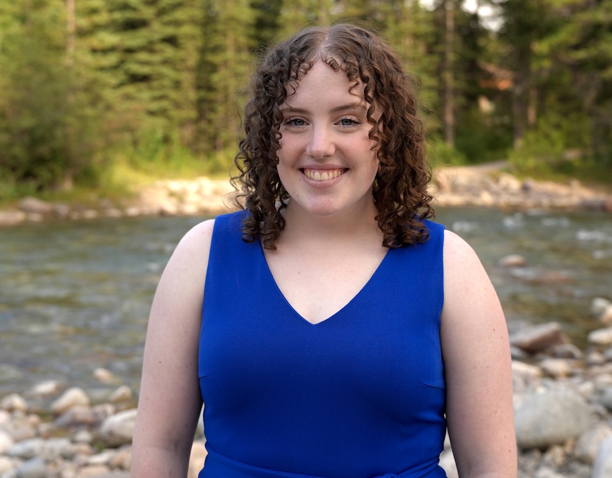 A woman with light skin tone and shoulder-length curly brown hair wears a blue V-neck dress.