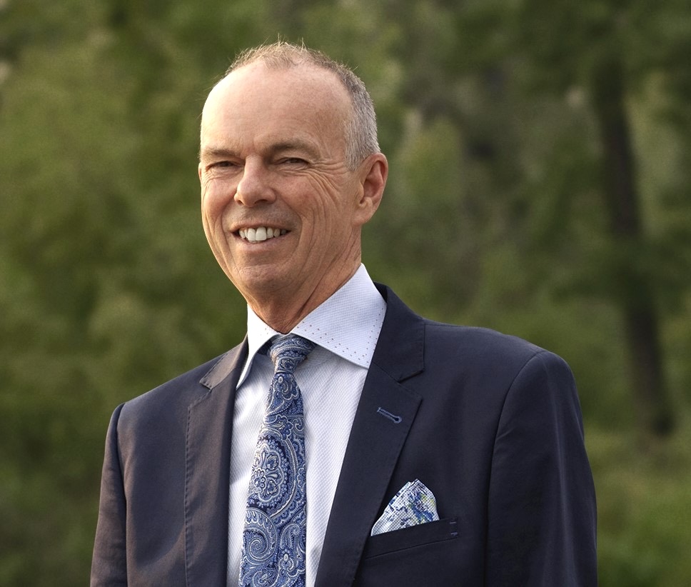 A man with close-cropped, balding hair and light skin tone wears a dark suit jacket and a paisley tie.
