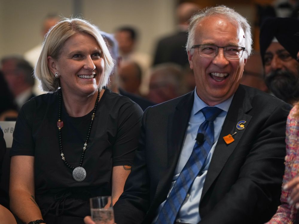 Elenore Sturko, a light-skinned woman with short blond hair, smiles as she sits beside a laughing John Rustad.