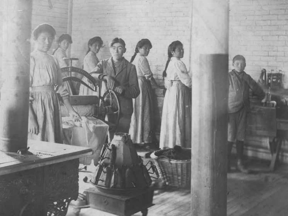 Seven Indigenous teens &mdash; two boys and five girls &mdash; pause while working in a laundry room to look at a photographer. 