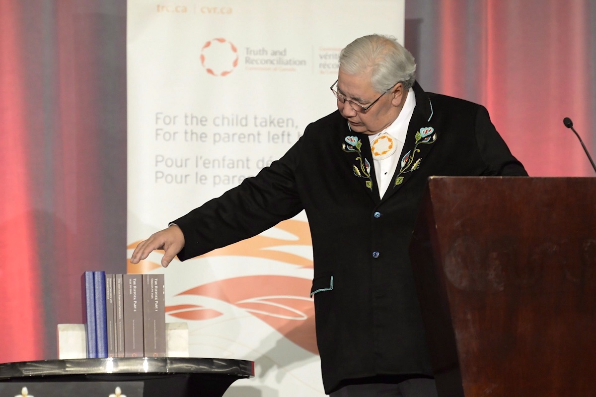 Justice Murray Sinclair is a grey-haired Indigenous man. He is wearing glasses and a formal black blazer, and moving his hands towards several books that comprise the final report of the Truth and Reconciliation Commission of Canada.