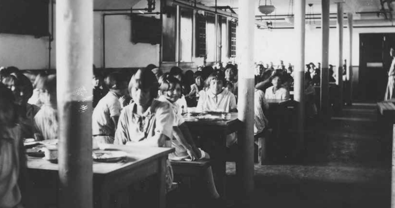 An interior shot of a large cafeteria room with students sitting at long, parallel tables.