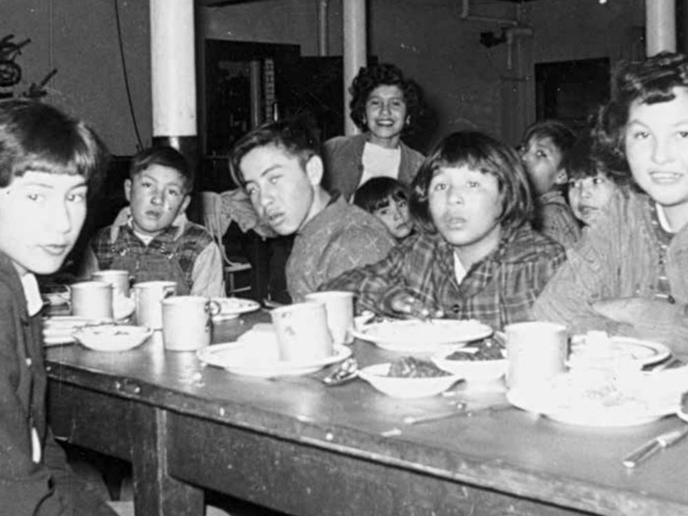A half-dozen residential school youth are seated along a narrow table crowded with small plates and cups.