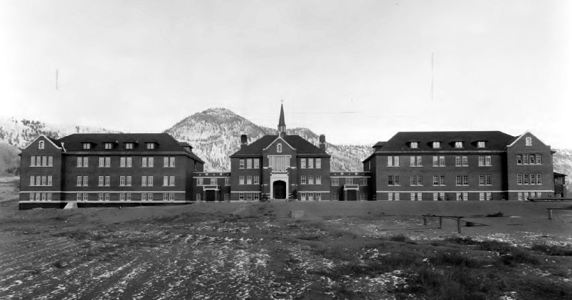The front view of the Kamloops Indian Residential School building, with a muddy terrain in the foreground and sparsely wooded hills in the background.