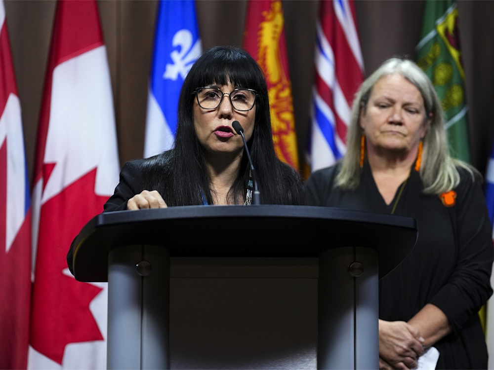 A woman with medium-light skin tone, straight black hair and wide glasses, member of Parliament Leah Gazan, is standing and speaking at a microphoned lectern. Behind her to the right is a woman with light skin tone and fair hair, special interlocutor Kimberly Murray, wearing orange earrings and a pin on her dress with an orange shirt on it.