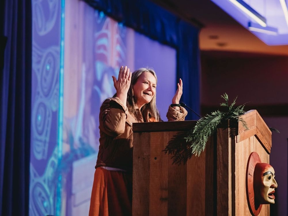 A middle-aged woman, independent special interlocutor Kimberly Murray, with fair and grey hair speaks onstage with her forearms up in front of a wooden stand in a dimly lit room. She is wearing a brown jacket and an orange skirt.